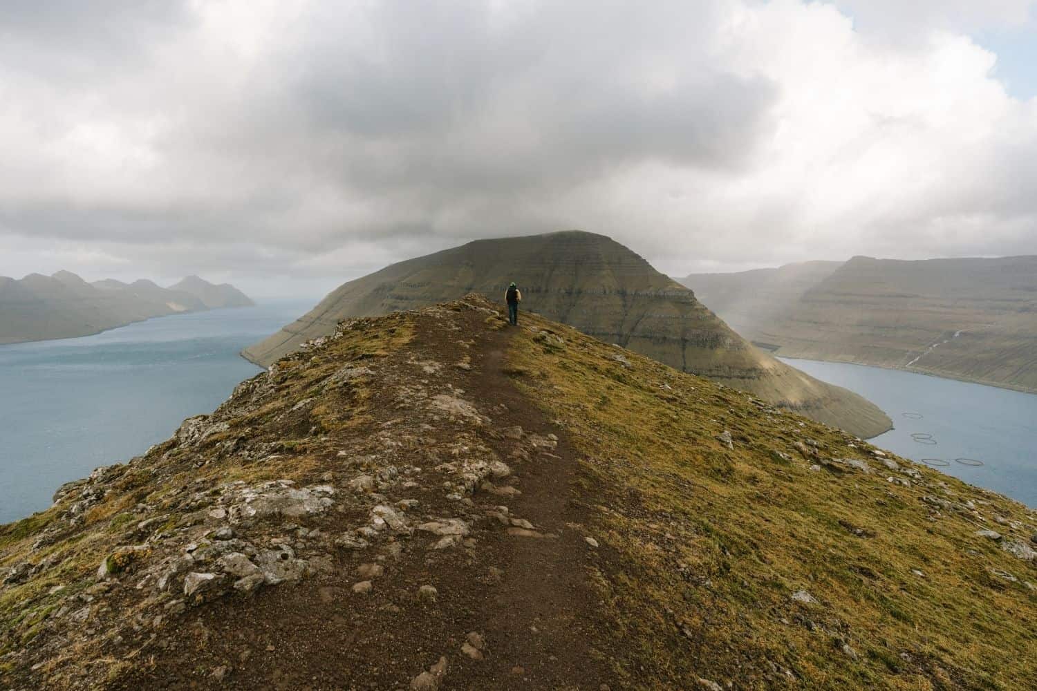 Hiker standing at the top of Klakkur with dramatic mountain and fjord views in the Faroe Islands.